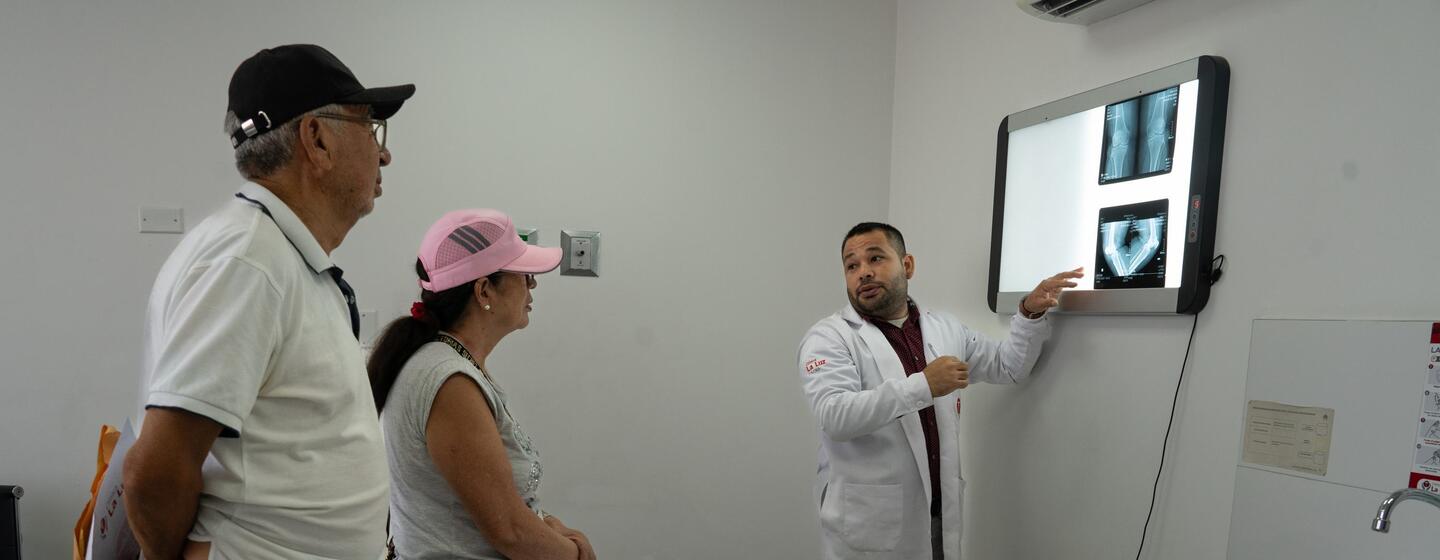 Un médico con bata blanca señala las radiografías en una pantalla mientras se las explica a un hombre y una mujer mayores en una sala de consulta.