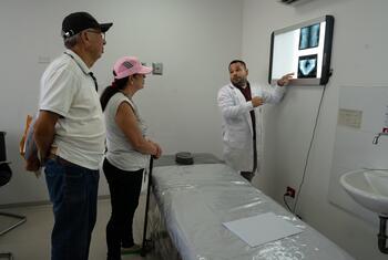 Un médico con bata blanca señala las radiografías en una pantalla mientras se las explica a un hombre y una mujer mayores en una sala de consulta.