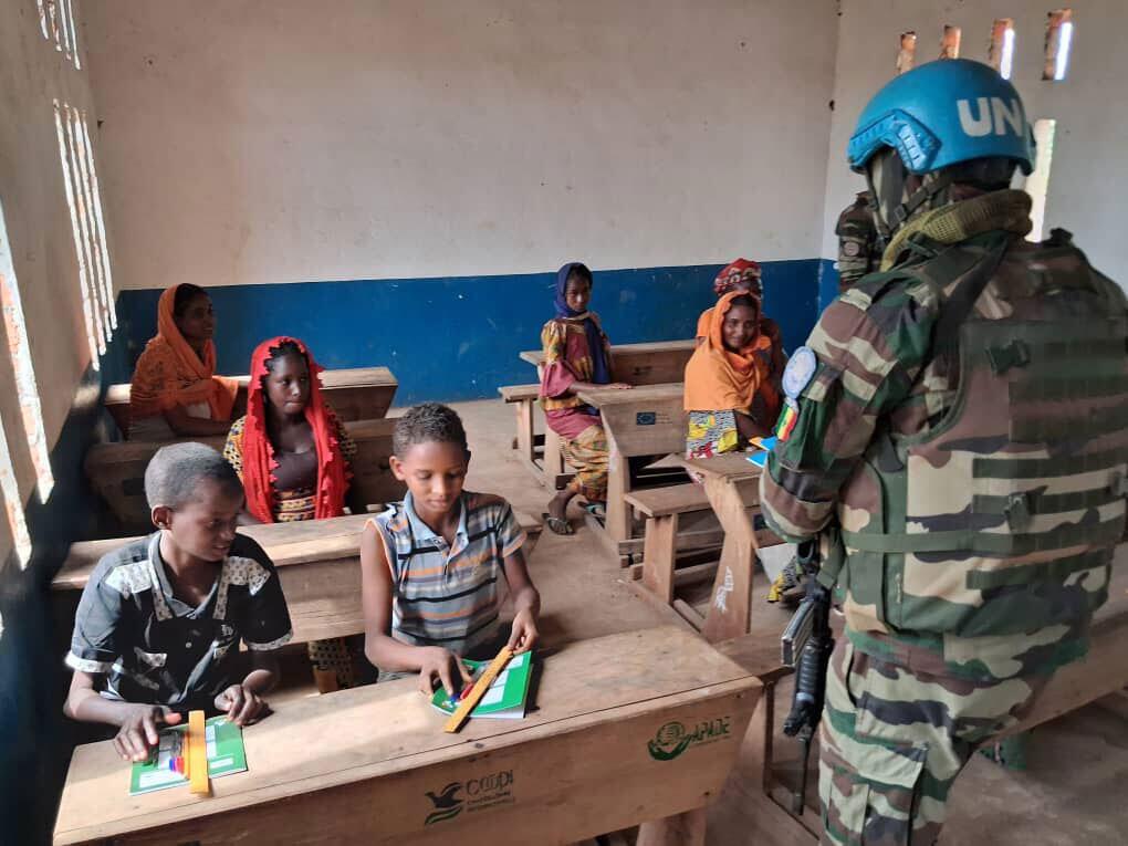 A UN peacekeeper in a blue helmet and camouflage uniform stands in a classroom with children.