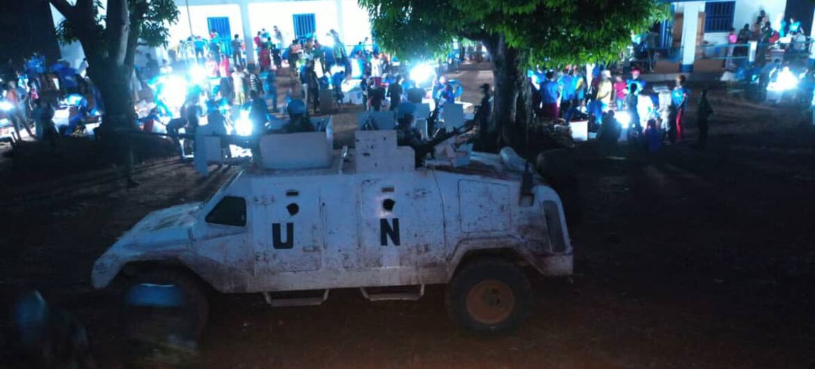 Un véhicule blindé de ONU est stationné de nuit, avec une foule de personnes rassemblées autour d'un bâtiment en arrière-plan.