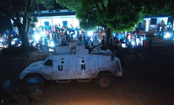 Un véhicule blindé de ONU est stationné de nuit, avec une foule de personnes rassemblées autour d'un bâtiment en arrière-plan.
