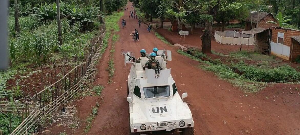 Des Casques bleus de ONU , à bord d'un véhicule blindé, circulent sur une route rurale de la République centrafricaine, avec un village et des palmiers en arrière-plan.