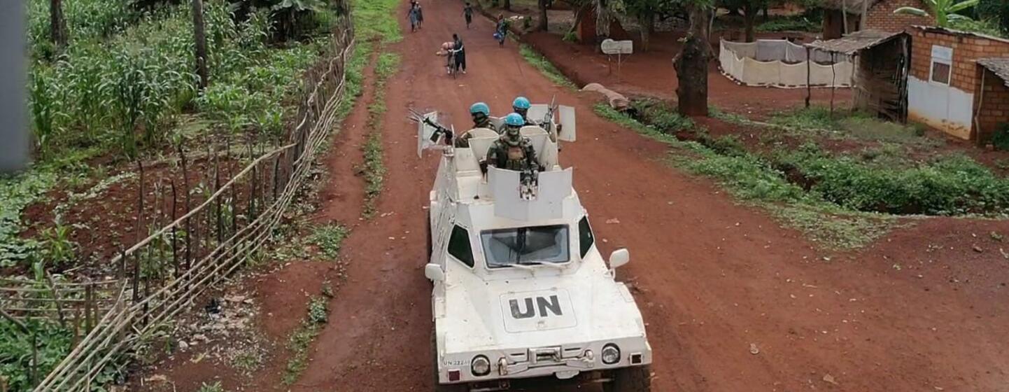 UN peacekeepers in an armored vehicle drive down a rural road in the Central African Republic, with a village and palm trees in the background.