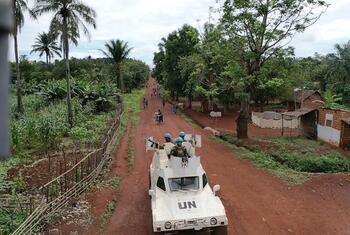 Des Casques bleus de ONU , à bord d'un véhicule blindé, circulent sur une route rurale de la République centrafricaine, avec un village et des palmiers en arrière-plan.