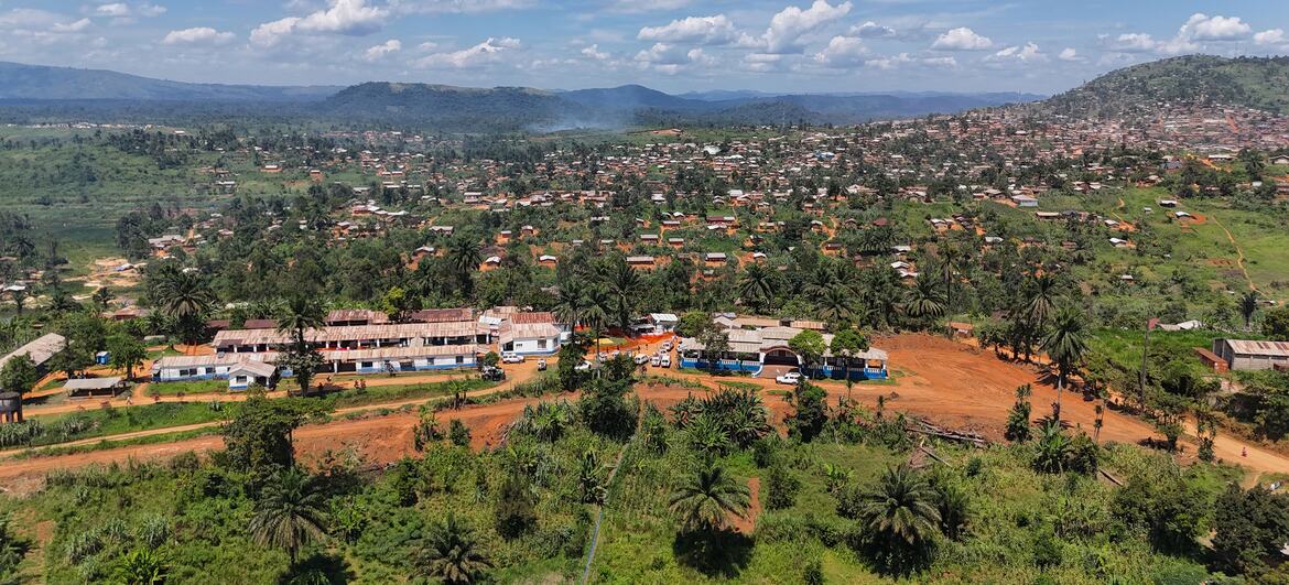 Aerial view of a rural town in DR Congo with a field hospital complex in the foreground, surrounded by green vegetation and hills under a cloudy sky.