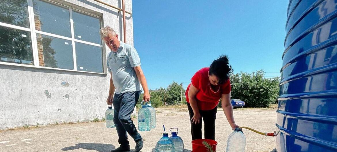 В Каховке, Украина, мужчина и женщина наполняют большие кувшины водой из синего резервуара во время проведения гуманитарной акции.
