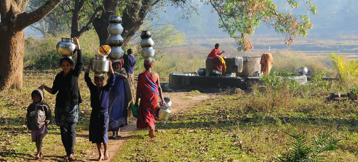 Women and children walk along a path carrying metal pots on their heads, with others at a water station in a rural setting.