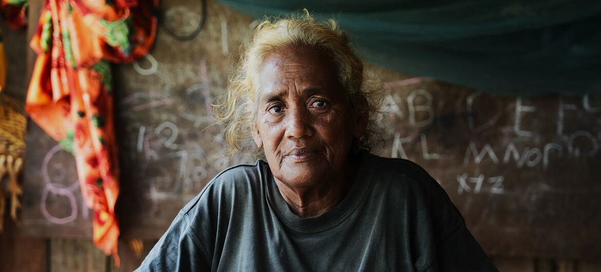 Portrait of an elderly woman with grey hair looking at the camera, wearing a dark t-shirt with an adidas logo. She is seated indoors in front of a wooden wall with a chalkboard that has numbers and letters written on it.