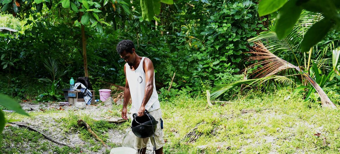 A man in a white tank top and shorts stands next to a well in a rural, tropical setting, pouring water from a black bucket into a white bucket.