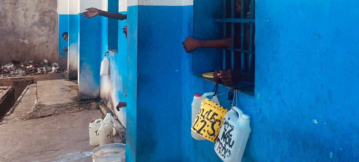 Se ven las manos de la gente a través de las ventanas enrejadas de un edificio azul y blanco, con garrafas de agua colgando en el exterior. Al fondo, sobre un suelo de cemento, se observa un montón de basura.