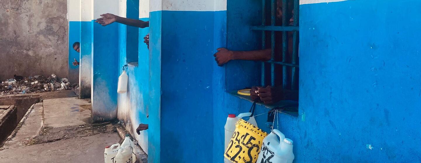 People's hands are visible through barred windows of a blue and white building, with water jugs hanging outside. A pile of trash is in the background on a concrete ground.