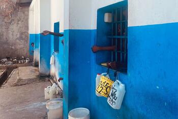 É possível ver as mãos das pessoas através das janelas gradeadas de um prédio azul e branco, com jarras de água penduradas do lado de fora. Ao fundo, vê-se uma pilha de lixo sobre o chão de concreto.