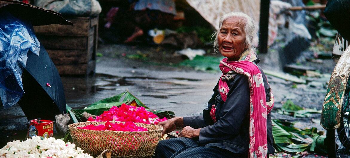 Una anciana está sentada en una calle mojada de Indonesia, vendiendo cestas llenas de pétalos de flores de un rojo y blanco vibrantes.