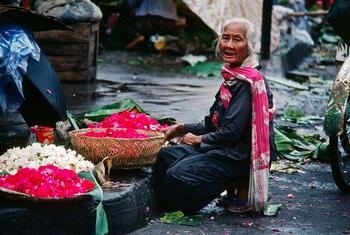 Una anciana está sentada en una calle mojada de Indonesia, vendiendo cestas llenas de pétalos de flores de un rojo y blanco vibrantes.