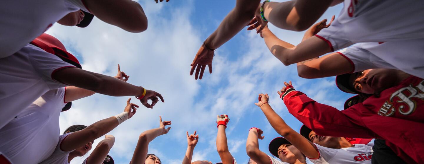 Equipo femenino de sóftbol reunido en círculo, visto desde abajo, bajo un cielo azul.