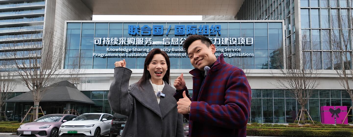 一名中国女子和一名中国男子在一座现代建筑前竖起大拇指合影，该建筑上有关于联合国可持续采购方案的标牌。