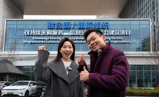一名中国女子和一名中国男子在一座现代建筑前竖起大拇指合影，该建筑上有关于联合国可持续采购方案的标牌。
