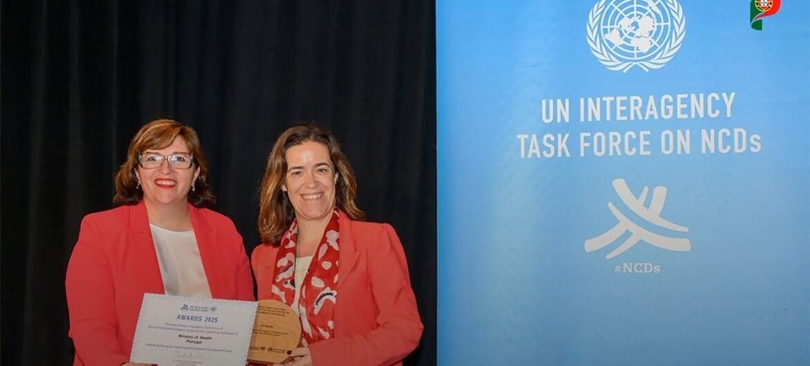 Duas mulheres de blazer vermelho posam em uma cerimônia de premiação, segurando um certificado e uma placa. Ao fundo, em azul, lê-se "Força-Tarefa Interagencial da ONU sobre Doenças Não Transmissíveis", com o logotipo da ONU e a bandeira portuguesa.