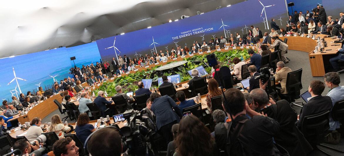 Delegados participam de uma sessão temática sobre transição energética na cúpula climática da UNFCCC, com uma grande mesa de conferência e imagens de turbinas eólicas ao fundo.