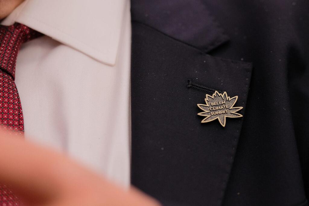 Primer plano de una persona que lleva una chaqueta de traje oscura, camisa blanca y corbata roja, con un broche dorado en la solapa que dice "BELEM CLIMATE SUMMIT".