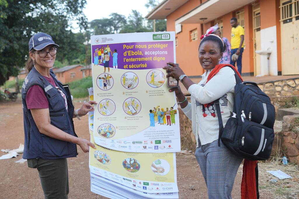 Mfanyakazi wa afya wa WHO na mwanamke wa eneo hilo wanashikilia bango kubwa la taarifa kuhusu kinga dhidi ya Ebola na desturi za mazishi zenye heshima huko Nyankunde, Ituri, DR Congo.