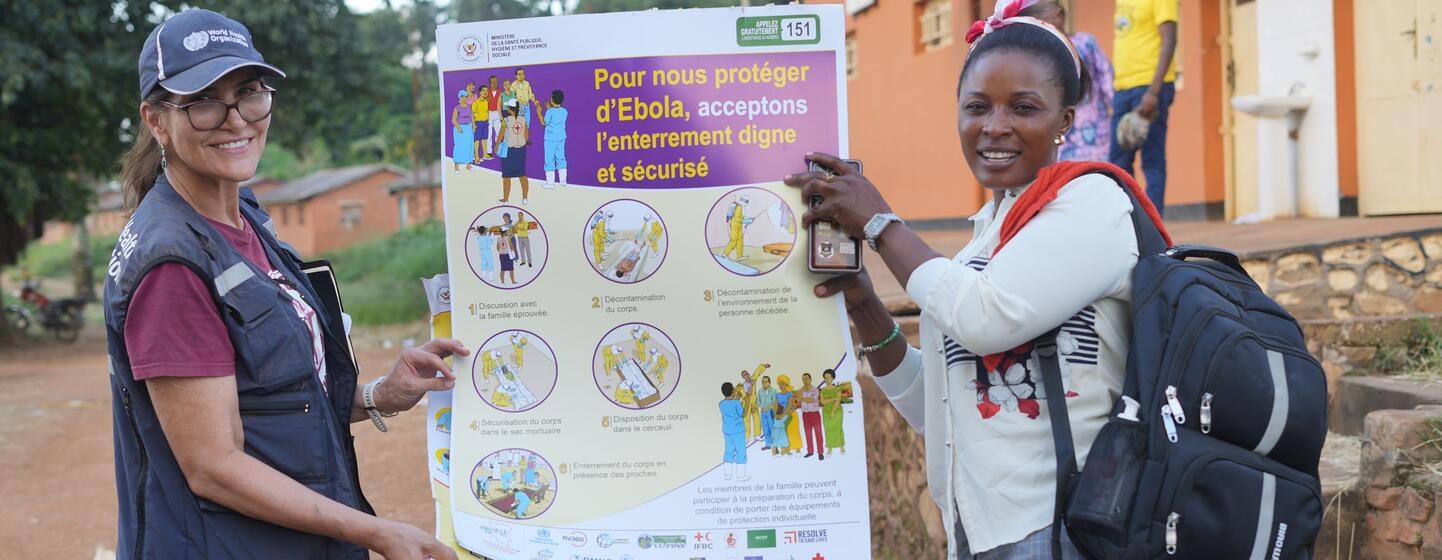 Un trabajador sanitario de la OMS y una mujer local sostienen un gran cartel informativo sobre la prevención del ébola y las prácticas de entierro digno en Nyankunde, Ituri, República Democrática del Congo.