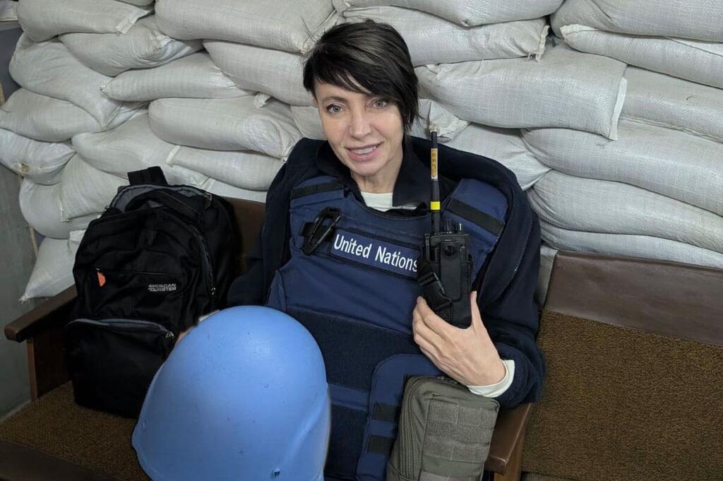 Una mujer, que trabaja en seguridad, lleva un chaleco de las Naciones Unidas y sostiene un walkie-talkie, sentada en una silla frente a un muro de sacos de arena.
