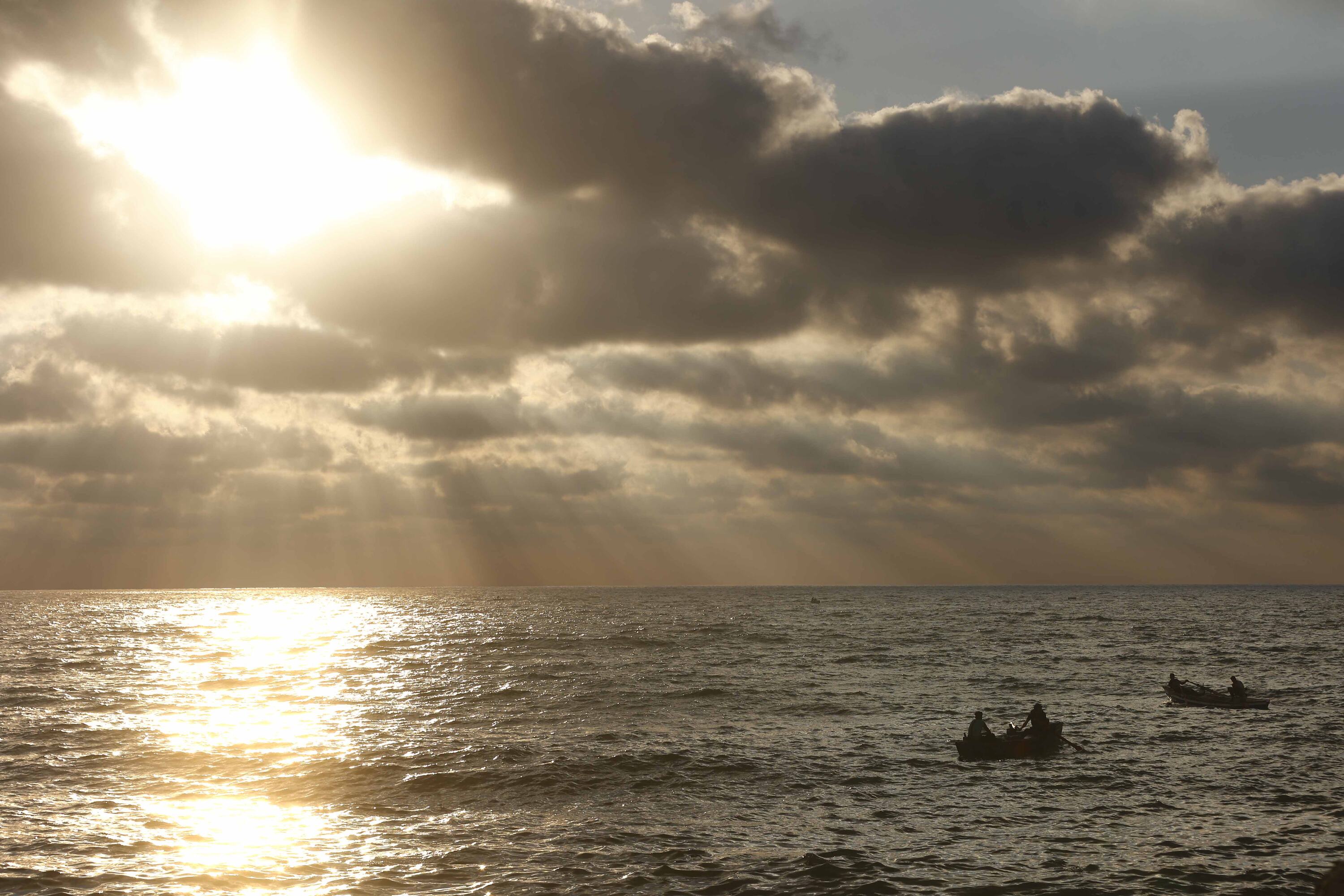 صور ظلية لقاربين للصيد مع صيادين على الماء، والشمس تغرب خلف سماء ملبدة بالغيوم.
