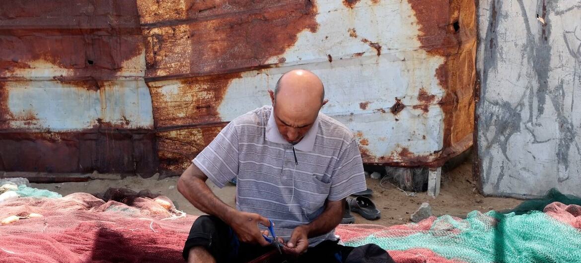 Un hombre calvo con una camisa de rayas está sentado sobre una gran red de pesca roja en el puerto de Gaza, usando unas tijeras para reparar la malla.