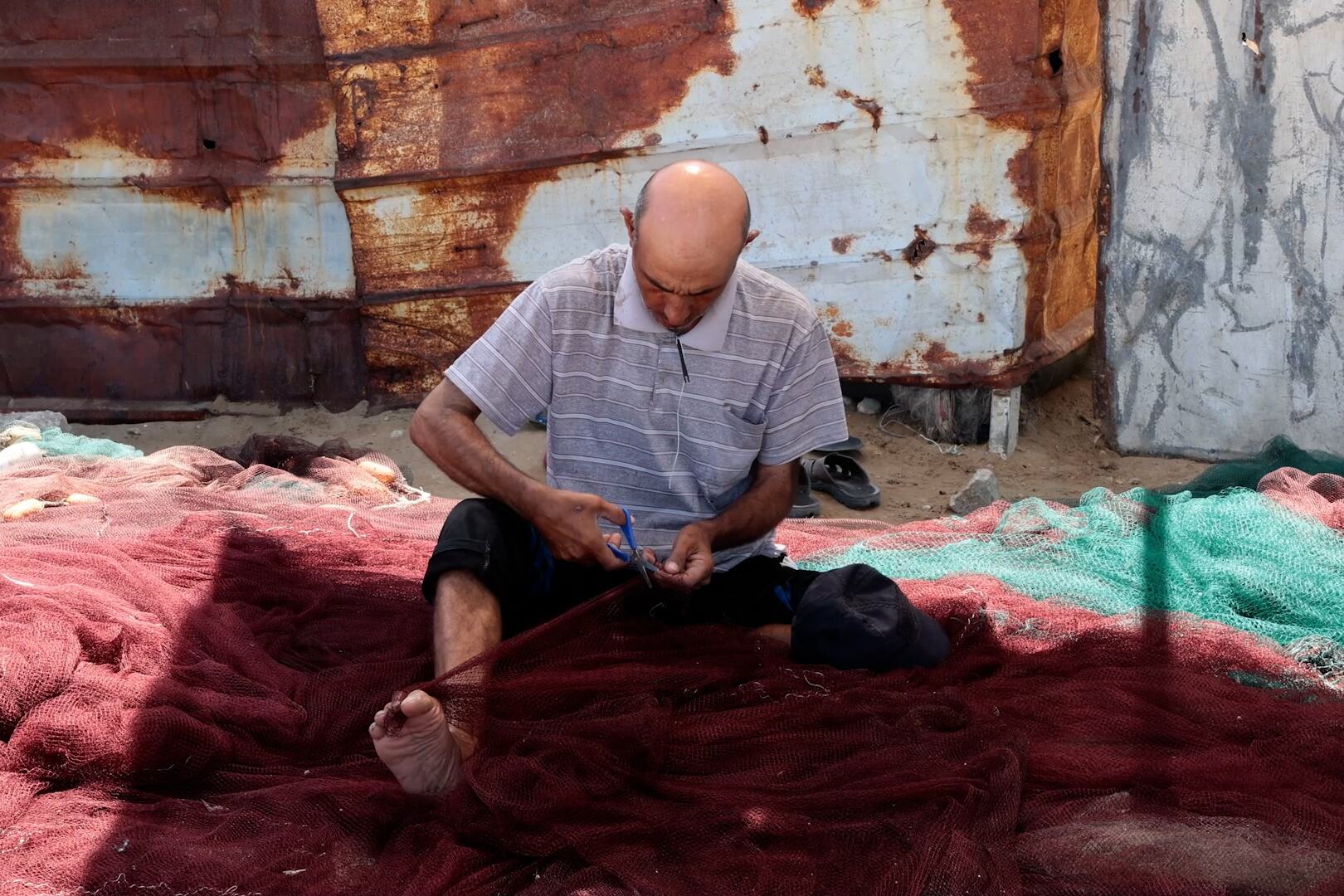 رجل أصلع يرتدي قميصاً مخططاً يجلس على شبكة صيد حمراء كبيرة في ميناء غزة، ويستخدم مقصاً لإصلاح الشبكة.