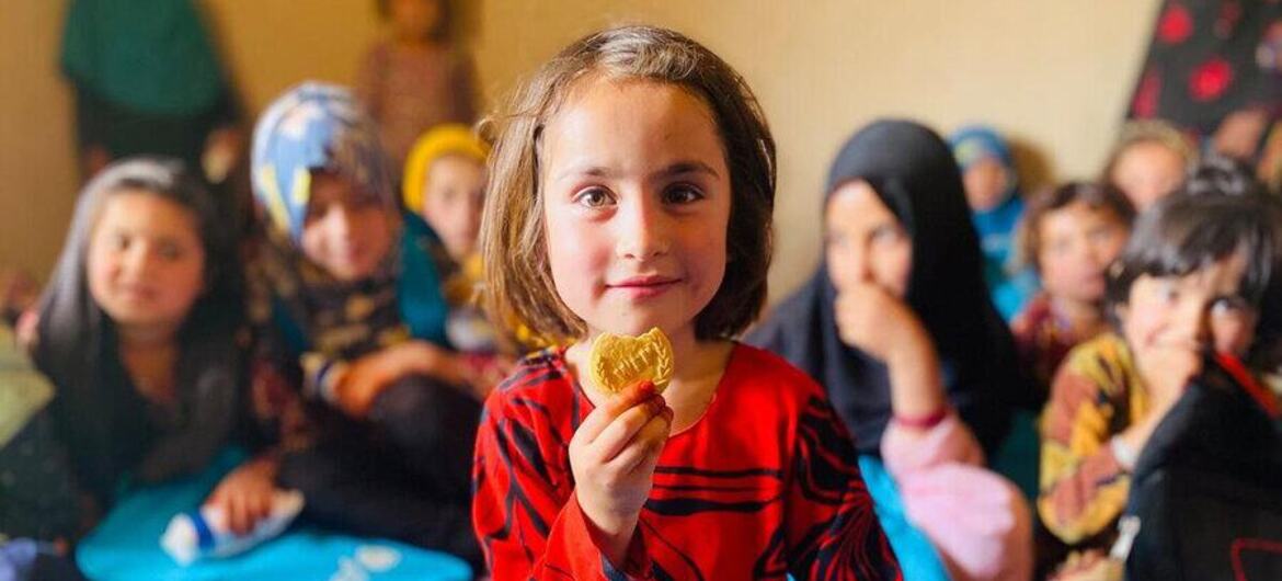 Una niña en un aula de Afganistán sostiene una galleta fortificada, con otros estudiantes al fondo. Esto forma parte del Programa de Alimentación Escolar del Programa Mundial de Alimentos en la provincia de Zabul.