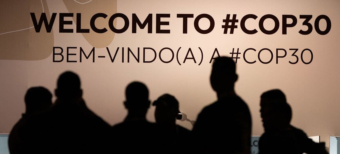 Silhouettes of people stand before a large sign that reads 'WELCOME TO #COP30' and 'BEM-VINDO(A) A #COP30' at the UNFCCC event in Belém.