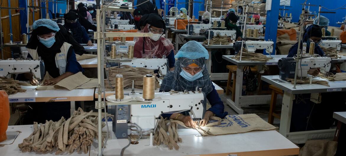 Mulheres refugiadas rohingya trabalham em máquinas de costura em uma fábrica de roupas em Bangladesh, com logotipos do ACNUR visíveis nos equipamentos.