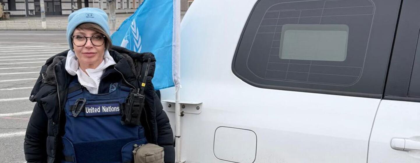 UN Security worker Olga Scripovaisa stands on a street in Odesa, Ukraine, next to a UN vehicle with the blue UN flag visible.