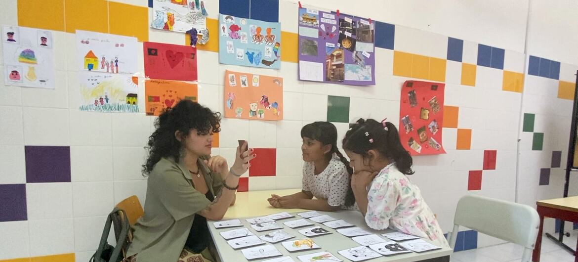 Uma mulher de cabelos cacheados ensina português a duas meninas sentadas à mesa em uma sala de aula em Lisboa, Portugal.