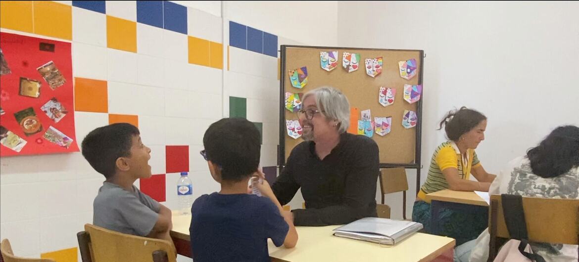 Uma professora está sentada em uma mesa conversando com dois jovens alunos do sexo masculino em uma sala de aula.