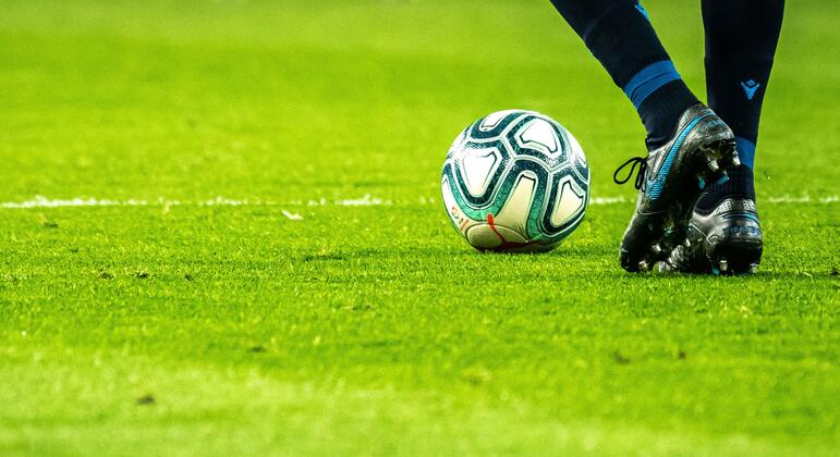 Primer plano de los pies de un jugador de fútbol con botas de fútbol junto a un balón en un campo verde.