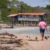 Two people walk along a sandy beach with driftwood in the foreground, towards a wooden stilt house on the shore, with a dense forest in the background.