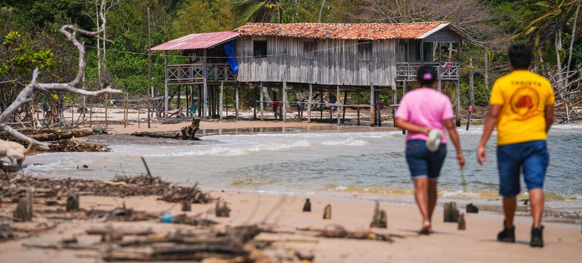 Deux personnes marchent le long d'une plage de sable fin, avec du bois flotté au premier plan, en direction d'une maison sur pilotis en bois sur le rivage, avec une forêt dense en arrière-plan.