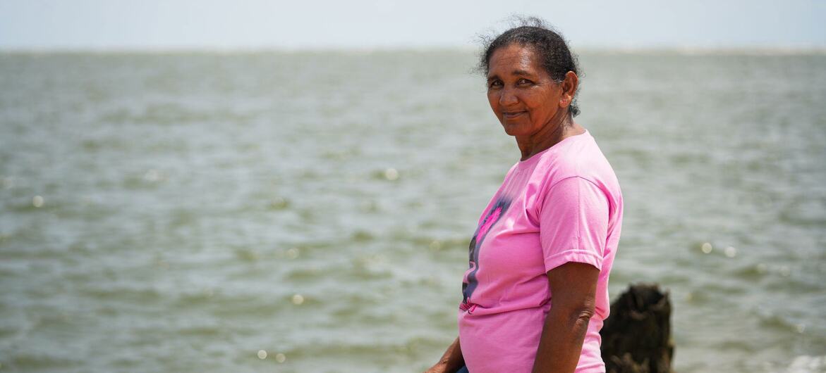 An elderly Brazilian woman in a pink shirt stands on a shoreline, looking back over her shoulder at the camera with the sea in the background.