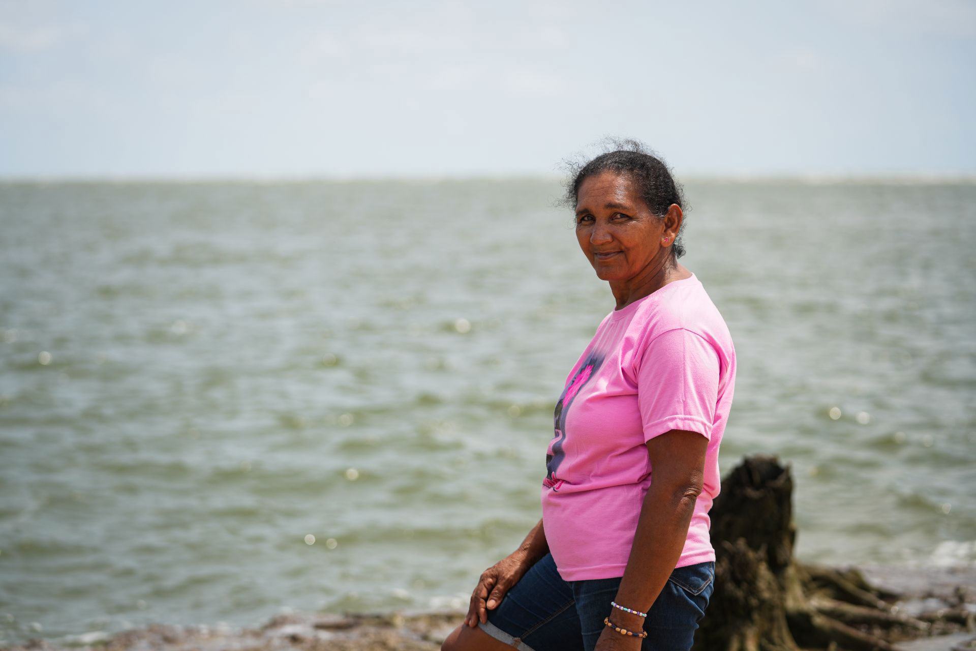 Une Brésilienne âgée, vêtue d'une chemise rose, se tient sur le rivage, regardant par-dessus son épaule vers la caméra, la mer en arrière-plan.
