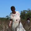 Uma senhora idosa, vestida com um vestido claro bordado com flores, colhe plantas em um campo seco no Brasil, enquanto outra pessoa trabalha ao fundo.