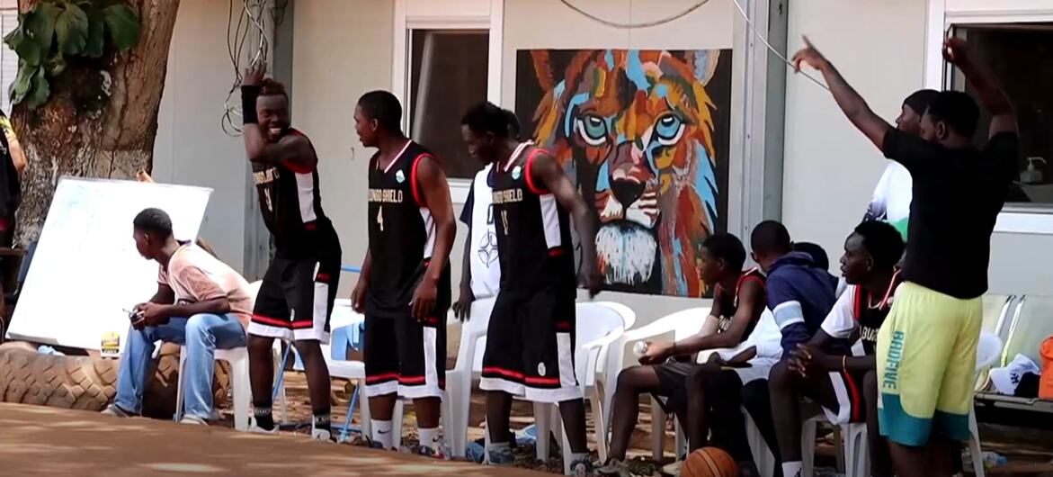 Kundi la wanaume wa Kiafrika wakiwa wamevaa sare za mpira wa kikapu na nguo za kawaida kwenye uwanja wa nje huko Yambio, wakiwa na ukuta wa simba wenye rangi nyingi nyuma.