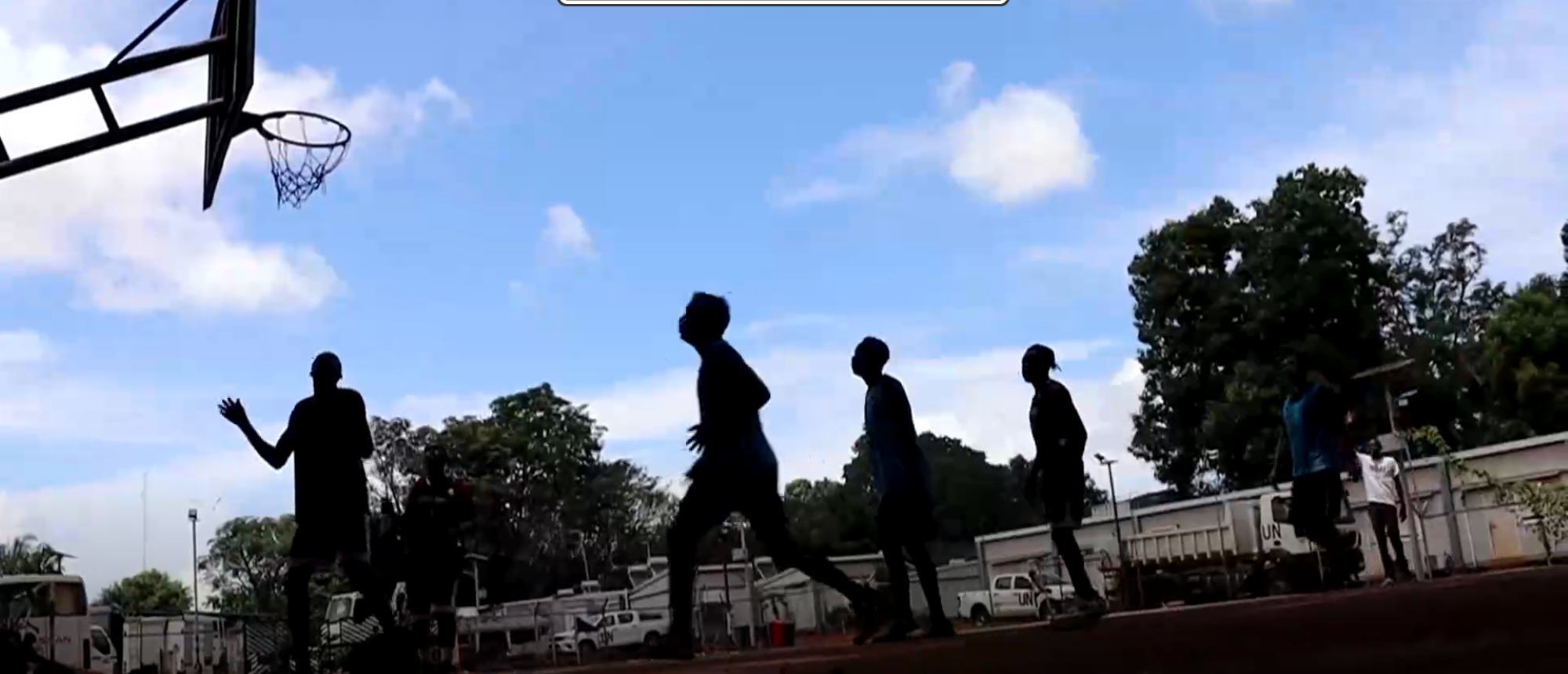 Silhouette za vijana wakicheza mpira wa kikapu kwenye uwanja wa nje chini ya anga la bluu, huku malori UN yakionekana nyuma.