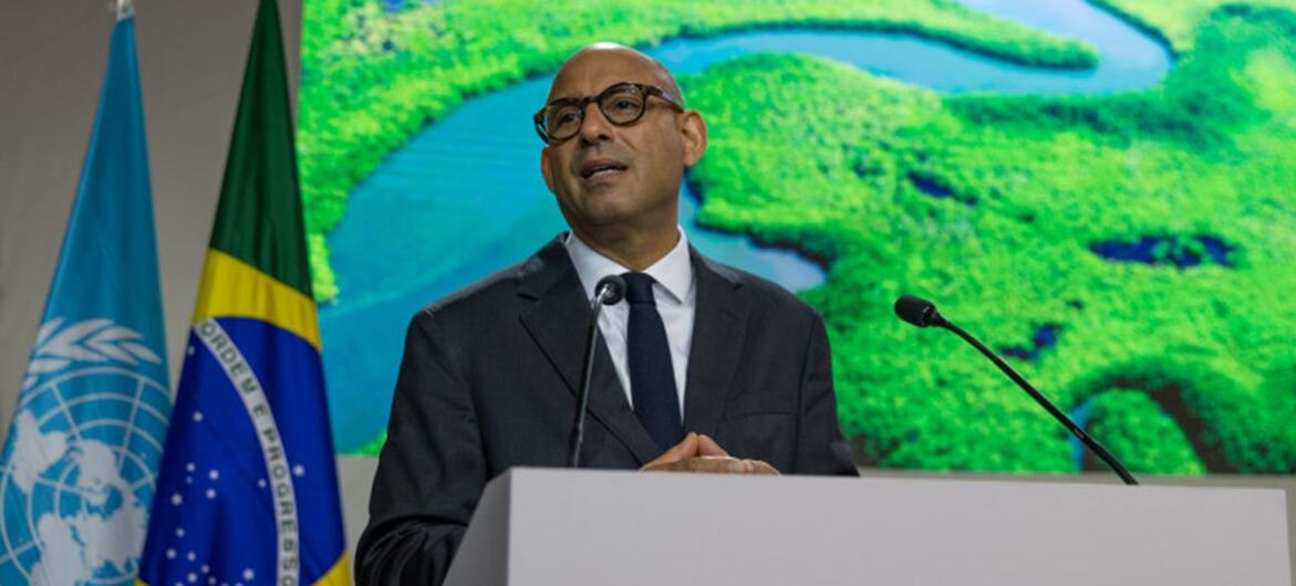 Um homem de terno e óculos discursa em um pódio na conferência COP30, com as bandeiras da ONU e do Brasil atrás dele.