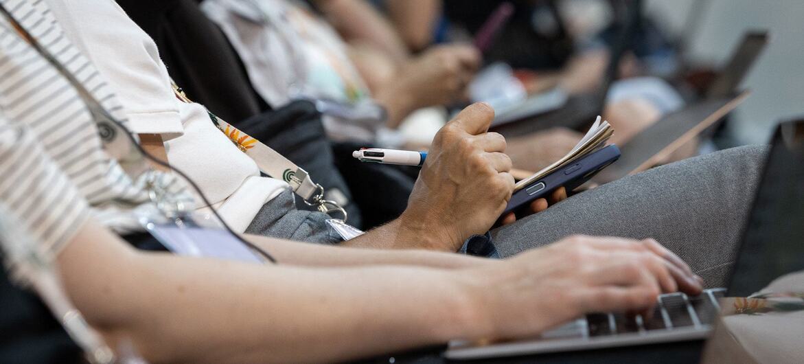 En la imagen se ve a los asistentes a una conferencia de perfil, con una persona tomando notas y otra usando un ordenador portátil.