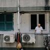 Un homme âgé en t-shirt blanc se tient sur un balcon à côté de climatiseurs et de linge étendu sur une corde à linge.