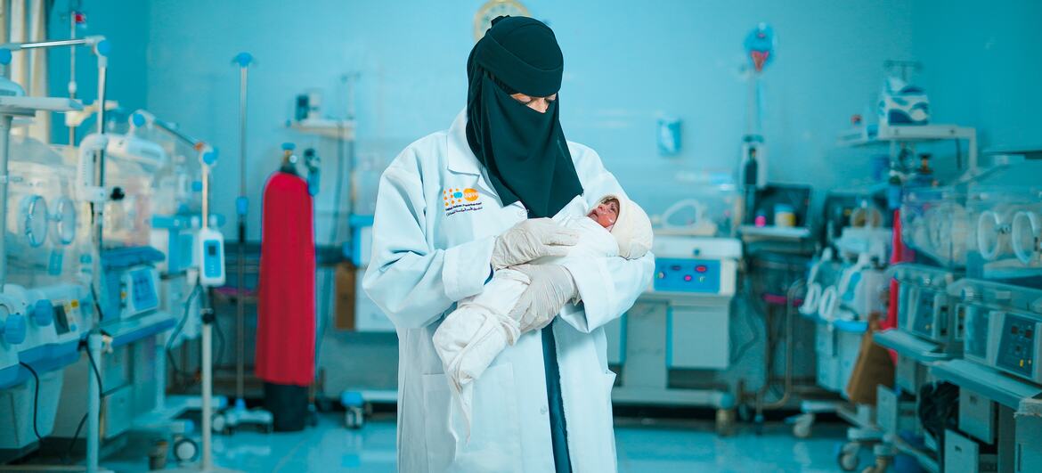 قابلة ترتدي معطفاً أبيض وحجاباً أسود تحمل طفلاً حديث الولادة في وحدة العناية المركزة لحديثي الولادة.