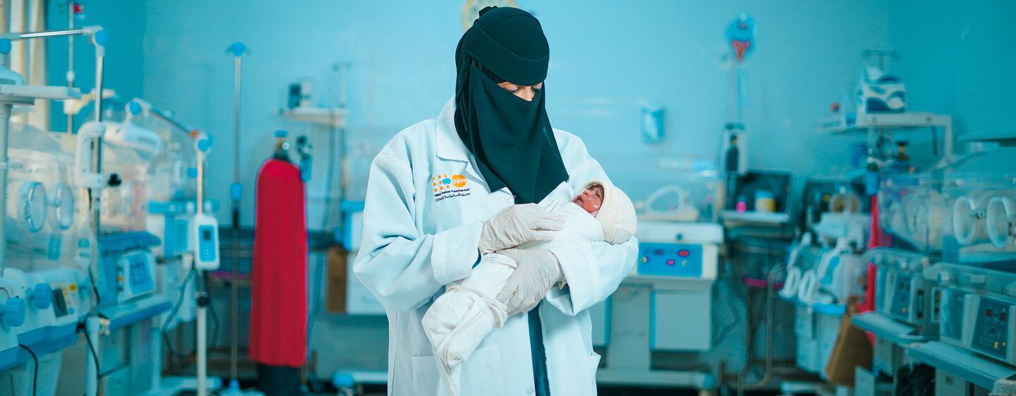 A midwife in a white coat and black hijab holds a newborn baby in a neonatal intensive care unit in Marib, Yemen.