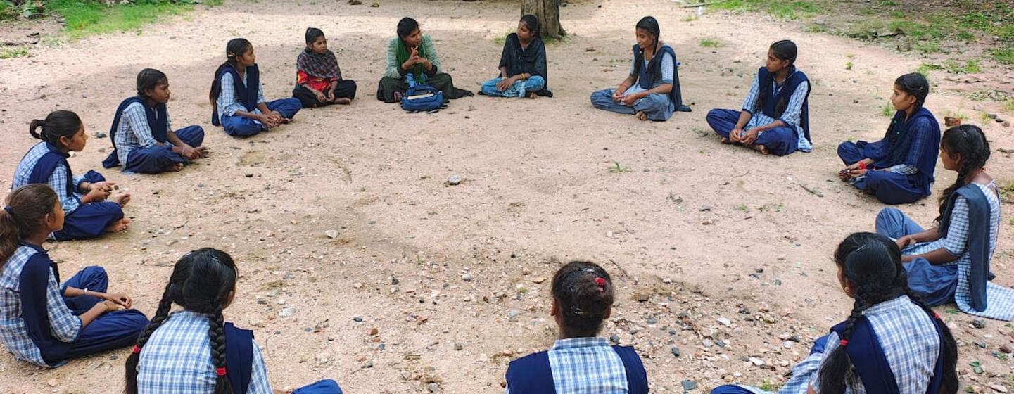नीली और सफेद वर्दी पहने भारतीय स्कूली छात्राओं का एक समूह दो वयस्क महिलाओं के साथ जमीन पर एक घेरे में बैठा है, जो संभवतः UNV की स्वयंसेवक हैं, एक बाहरी सत्र के दौरान।